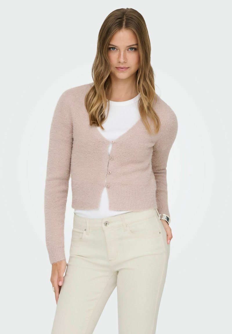 Young woman with loose wavy hair wears a light pink cropped cardigan over a white top and cream-colored pants against a plain background.