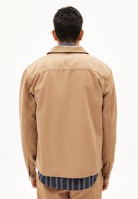 Tan buitenjack met een kraag, stevige stof, met knoopmanchetten en een rechte snit; gelaagd over een blauwe gestreepte blouse daaronder.