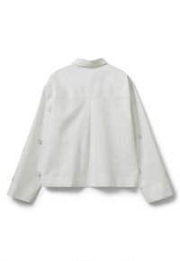 Hvid, løstsiddende skjorte med lange ærmer og krave. Har dekorative mønstre nær manchetterne og en plissering detalje på ryggen.