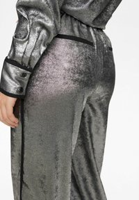 Metallic pilkos spalvos kelnės su minkšta tekstūra, turinčios juodą juosmens juostą ir šoninius apdailos elementus. Medžiaga turi šiek tiek blizgesio ir lygią paviršių.