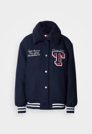 Jachetă varsity albastru marin cu guler din blană artificială, manșete cu dungi albe, logo-uri brodate și închidere cu capse. Dispune de buzunare laterale.