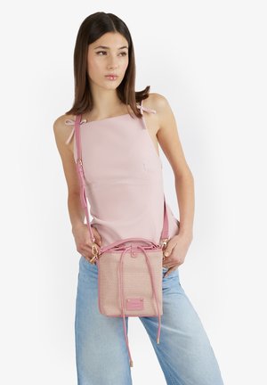 Donna che indossa un top senza maniche rosa chiaro e jeans blu, che tiene una borsa a tracolla rosa intrecciata con chiusura a coulisse.