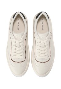 Ett par vita lädersneakers med vågformad brun kant, vita snören, svarta hälaccentuerar och "Cole Haan" tryckt inuti.