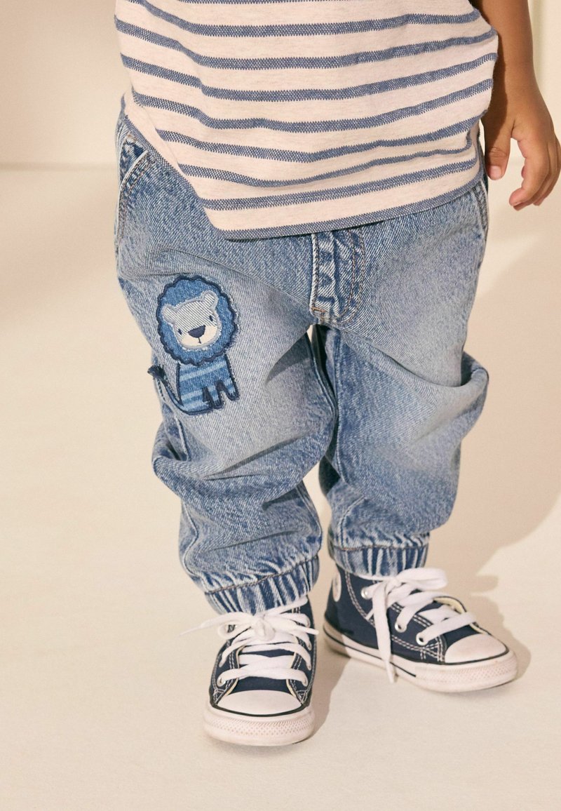 Barn iført blå jeans med en løveapplikation på benet, en stribet skjorte og marineblå sneakers, står på et lyst gulv.