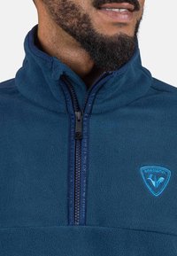 Felpa in pile blu navy con colletto alto, presenta una chiusura a zip a un quarto sul davanti e un logo ricamato blu sulla parte alta sinistra. Texture morbida, design casual.