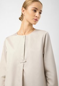 Beige halenka s dlouhým rukávem a kulatým výstřihem, se zapínáním na přední straně a dvěma knoflíky. Hladký materiál s volným střihem.