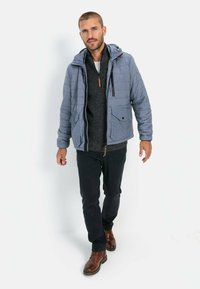 Lys blå polstret jakke med hætte, to forlommer og sort lynlås. Båret over en mørkegrå sweater og parret med mørke bukser og brune sko.