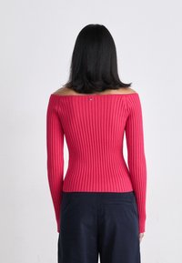 Maglione rosa a spalle scoperte, a coste, con maniche lunghe e un design aderente. Dettaglio notevole: piccolo logo sulla parte posteriore del collo. Visibile un paio di pantaloni blu navy.