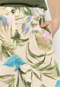 Floral linen pants with a creamy background, featuring green, blue, pink leaves, pleats, and a gold button at the waistband. Pockets visible.
