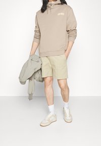 Felpa beige con cordino, logo sul petto, abbinata a pantaloni corti beige chiaro. Il modello tiene una giacca grigia, indossando calze bianche e sneakers.