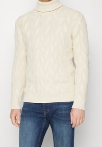 Cremfarbener Rollkragenpullover mit einem Zopfstrickmuster, gerippten Bündchen und Saum sowie einer strukturierten Oberfläche. Kombiniert mit blauen Jeans.