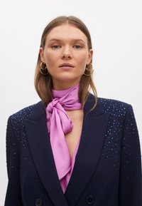 Blazer blu navy con spalle strutturate, decorato con paillettes blu sulla spalla sinistra, abbinato a una sciarpa di raso rosa annodata al collo.