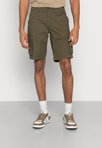 Pantaloni cargo verde oliva con due tasche laterali, vestibilità regolare. Tessuto in cotone, lunghezza fino a metà coscia, abbinati a scarpe da ginnastica marroni e calzini bianchi.