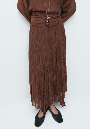 Brązowy, pognieciony, długi zestaw spódnicy i topu noszony z czarnymi, marszczonymi balerinami oraz złotym naszyjnikiem z czerwonym, łezkowatym wisiorkiem.