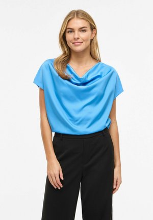 Kvinde med blondt hår iført en lyseblå draperet satinbluse og sorte skræddersyede bukser, stående mod en hvid baggrund.