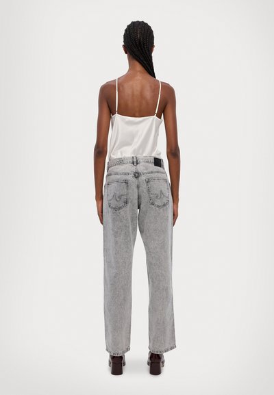 Pantalones vaqueros grises de cintura alta con un corte suelto, y una etiqueta negra en la parte trasera, combinados con una blusa de satén blanca y zapatos de tacón marrones.