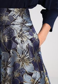 Hame floral dalam warna navy, yang menampilkan aksen biru muda dan emas. Terbuat dari kain tekstur dengan bentuk melebar dan memiliki saku samping.