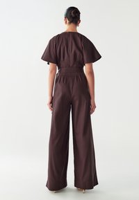 Mono marrón con piernas anchas, mangas cortas y ligeras, cintura entallada y detalle de cremallera en la parte posterior. Fabricado con una tela ligera y texturizada.