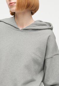 Γυναίκα με κοντά κόκκινα μαλλιά που φοράει ανοιχτό γκρι φούτερ Calvin Klein, το οποίο είναι κομμένο στο ύψος του πηγουνιού και δείχνει το άνω μέρος του κορμού και τις λεπτομέρειες της κουκούλας.