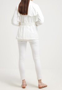 Vit bältesjacka med en flytande baksidesdesign, i kombination med vita smala jeans och lätta sandaler. Slät tyg med skräddarsydd passform.