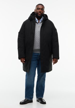 Homme portant un manteau d'hiver noir à capuche par-dessus un pull gris clair, un jean bleu et des chaussures noires, debout devant un fond blanc.