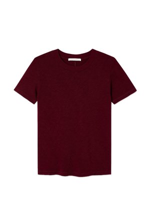 Personne debout contre un mur uni portant une chemise bordeaux à manches courtes, un jean bleu clair et des chaussures marron à lacets.