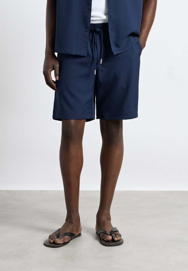 Homme portant un short bleu marine, une chemise assortie, un sous-maillot blanc et des sandales noires, debout sur un sol gris devant un fond blanc.