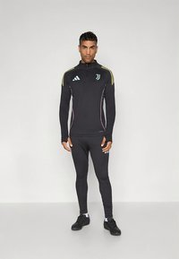 Musta urheilutakki ja -housut, joissa keltaiset ja vaaleansiniset yksityiskohdat, vetoketjullinen kaulus ja Adidas-logo rinnassa.