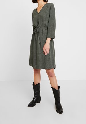 Frau trägt ein knielanges dunkelgrünes Kleid mit kleinen weißen Punkten und schwarze Lederstiefel bis zur Mitte der Waden, steht vor einem schlichten hellen Hintergrund.