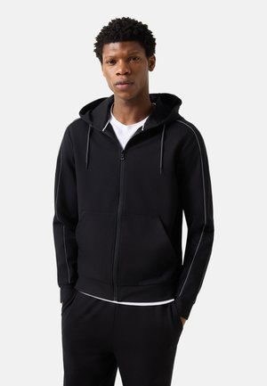 Junger Mann trägt schwarzen Zip-Hoodie mit weißen Paspeln und Hände in den Taschen, steht vor einfachem hellem Hintergrund.