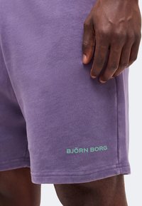 Κοντινή λήψη ενός σκούρου χρώματος χεριού που ακουμπά δίπλα σε λιλά σορτς με την επιγραφή «BJÖRN BORG» σε ανοιχτό πράσινο κοντά στο τελείωμα.
