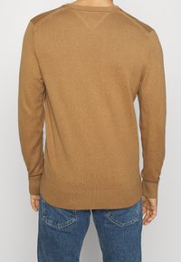 Ruskea neule, joka on valmistettu pehmeästä kankaasta. Siinä on röpelöinen pääntie ja hihansuut, ja sen muotoilu on vartalonmyötäinen, takana sileä rakenne.