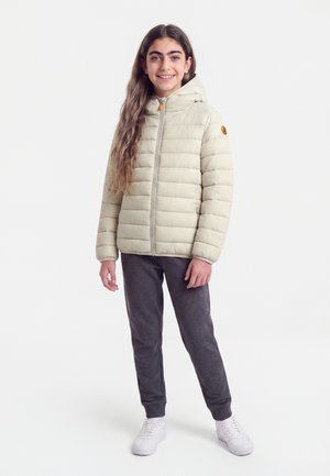 Teenagepige der smiler, iført en cremefarvet dynejakke med hætte, mørkegrå bukser og hvide sneakers, stående mod en ensfarvet baggrund.