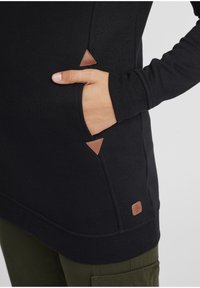 Fekete, hosszú ujjú tunika texturált anyagból, oldalzsebbel, háromszög alakú barna bőr kiegészítőkkel és egy kis márkajelzéssel.