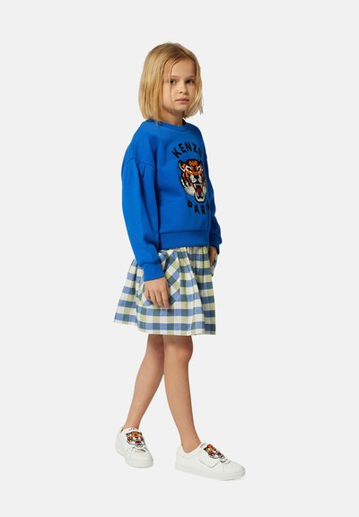 Sweatshirt bleu avec broderie de tigre, jupe à carreaux dans des tons de vert et de bleu, et baskets blanches avec détail de tigre. Tissu doux, coupe décontractée.