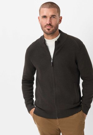 Man met kort haar en baard, draagt een donker vest met rits over een wit overhemd en bruine broek, handen in de zakken, kijkt vooruit.