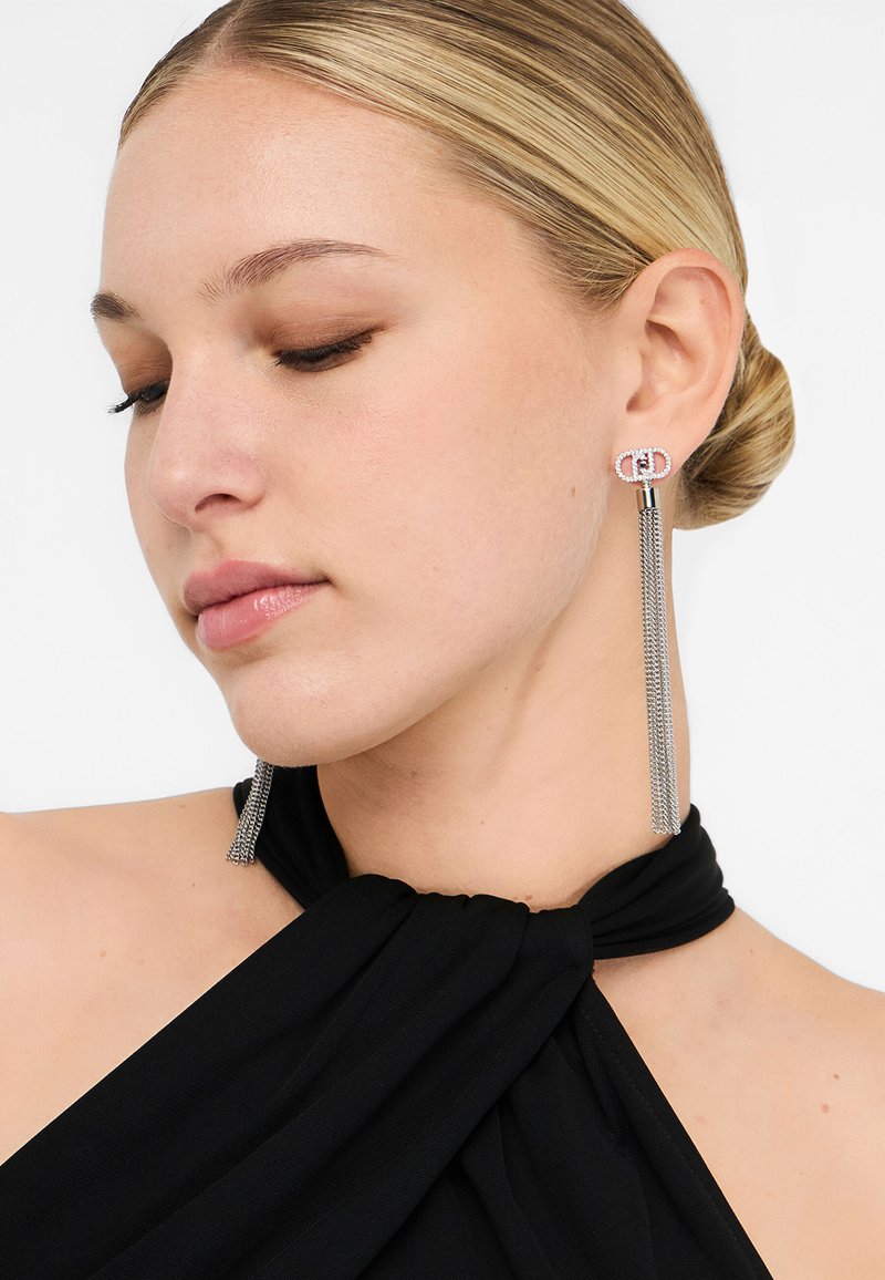 Ezüst bojtos fülbevalók kettős kör alakú díszítéssel, gyöngyökkel díszítve, hengeres szárból lelógva. Fekete ruhadarab háttere.