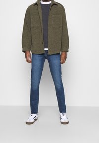 Grön texturerad skjorta med knappar över en grå tröja, i kombination med blå jeans och vita sneakers med svarta ränder. Två skjortfickor synliga.