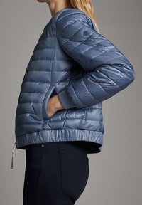 Personne en doudoune bleu clair et pantalon foncé, vue de côté avec les mains dans les poches sur un fond neutre.