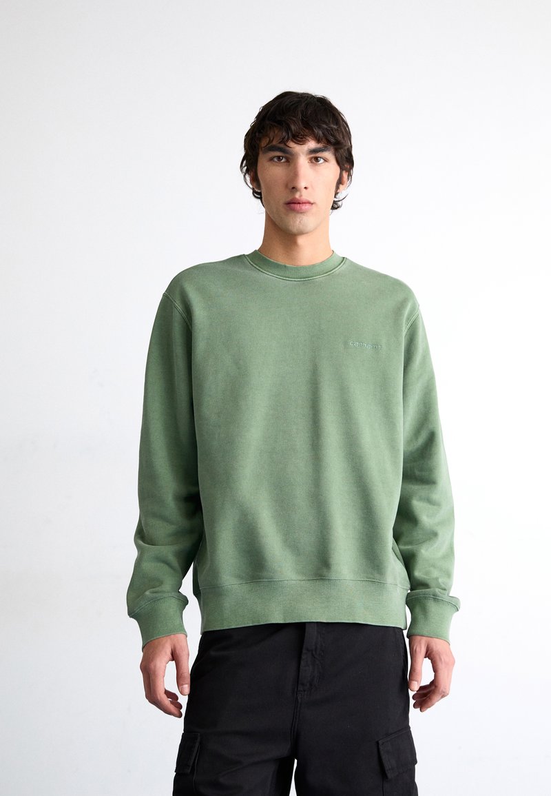 Grüner Pullover aus weichem Stoff, mit langen Ärmeln, rundem Hals und einem dezenten geprägten Logo auf der Brust, kombiniert mit schwarzen Hosen.