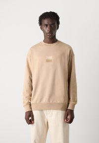 Beige sweatshirt in soft fabric, featuring a square logo in the center. Crew neck and ribbed cuffs add structure. Paired with light pants.