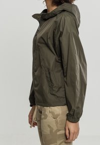 Olivgrön lättviktsjacka med huva, dragkedjestängning, elastiska ärmslut och sidofickor, kombinerad med beige kamouflagedelar. Slät, vattenavvisande material.