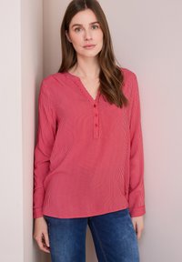 Maglietta a maniche lunghe a righe rosse e bianche con scollo a V, dotata di una chiusura con bottoni e una vestibilità comoda. Abbinata a jeans blu scuro.