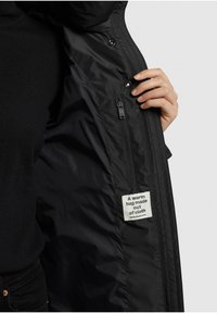 Černá prošívaná bunda s kapsou na zip a štítkem s nápisem "Teplé objetí vyrobené z látky." Hladký povrch a přiléhavý střih.