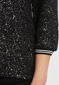 Schwarzer Pullover mit strukturierter Oberfläche und silbernen Sprenkeln, sowie einem gestreiften Rippbündchen. Das Material ist weich und hat einen lockeren Schnitt.