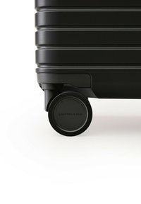 Zwarte hard-shell koffer met een gestructureerde afwerking, uitgerust met een opvallend wiel gemarkeerd met "KAPTEN & SON" voor soepele mobiliteit.
