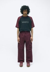 Bordeauxrode oversized broeken met zakken, gecombineerd met een zwart en bordeauxraglan T-shirt met een wit logo. Draag met bordeauxrode schoenen.