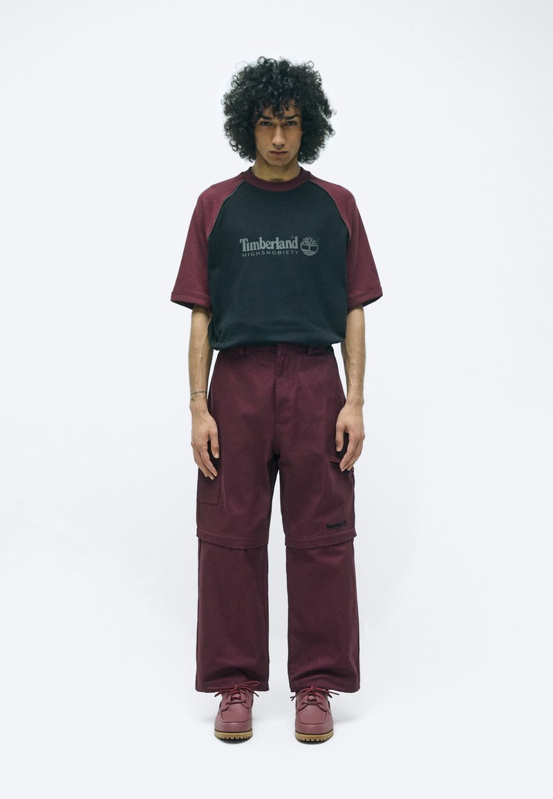 Bordeauxrode oversized broeken met zakken, gecombineerd met een zwart en bordeauxraglan T-shirt met een wit logo. Draag met bordeauxrode schoenen.