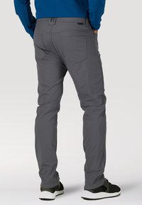 Mann trägt graue Cargohose und schwarze Sneakers, steht mit den Händen in den Taschen und ist mit dem Rücken vor einem einfarbigen Hintergrund zu sehen.