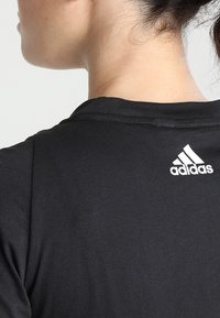 Černé bavlněné tričko s kulatým výstřihem. Na zadní straně je bílé logo Adidas. Krátké rukávy a hladká textura.